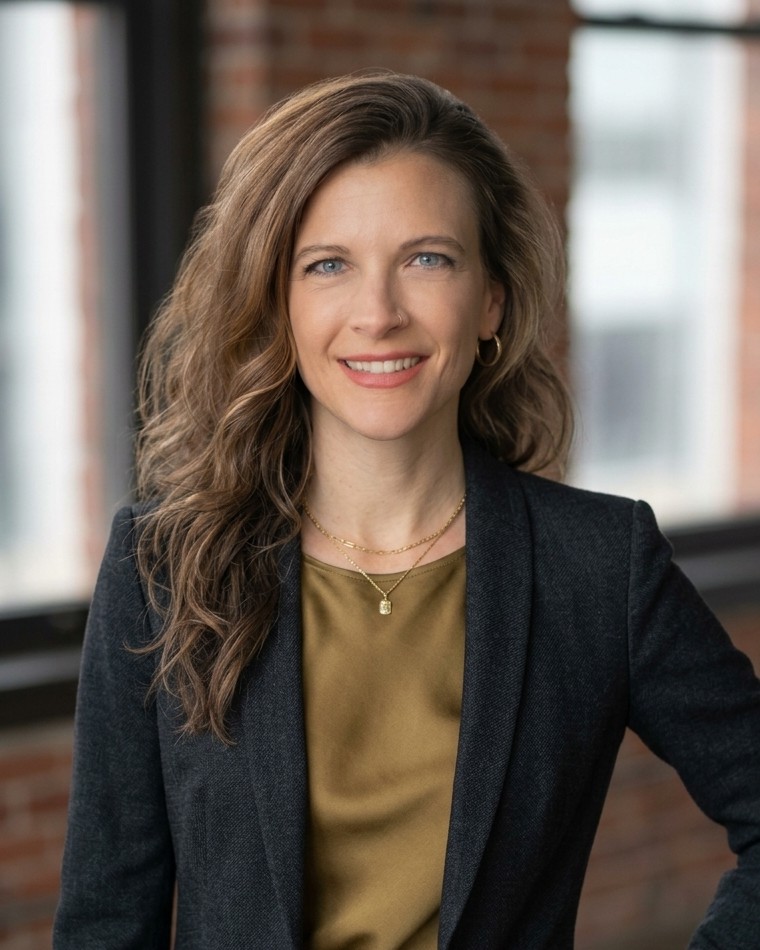 Amy Price, wearing a charcoal blazer over an olive top, smiling in a brick-walled office